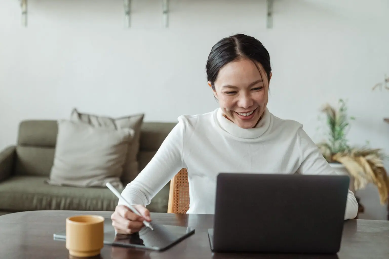 Female worker smiling at laptop while taking notes on tablet using digital pencil.